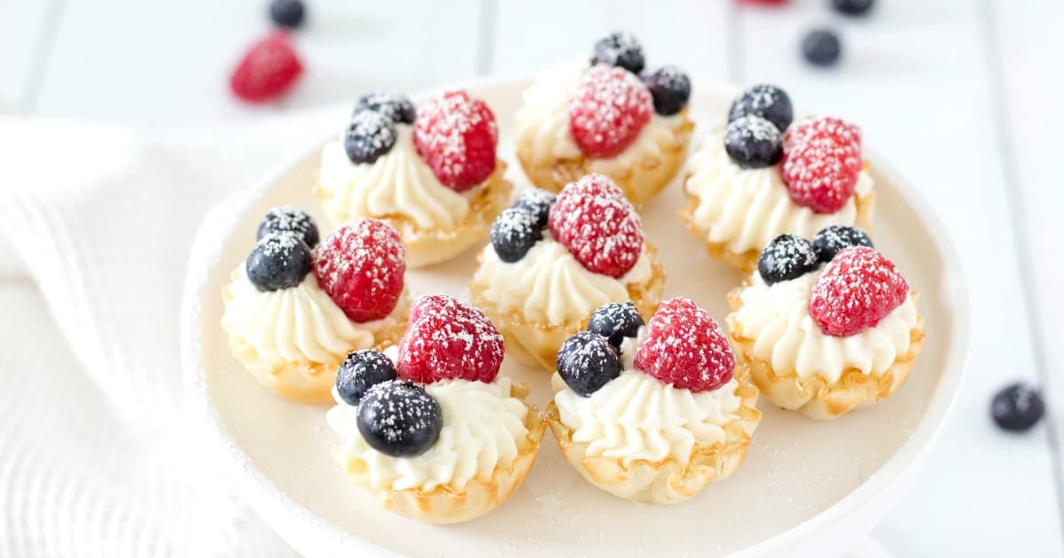 Mini tart desserts topped with whipped cream, raspberries, and blueberries dusted with powdered sugar
